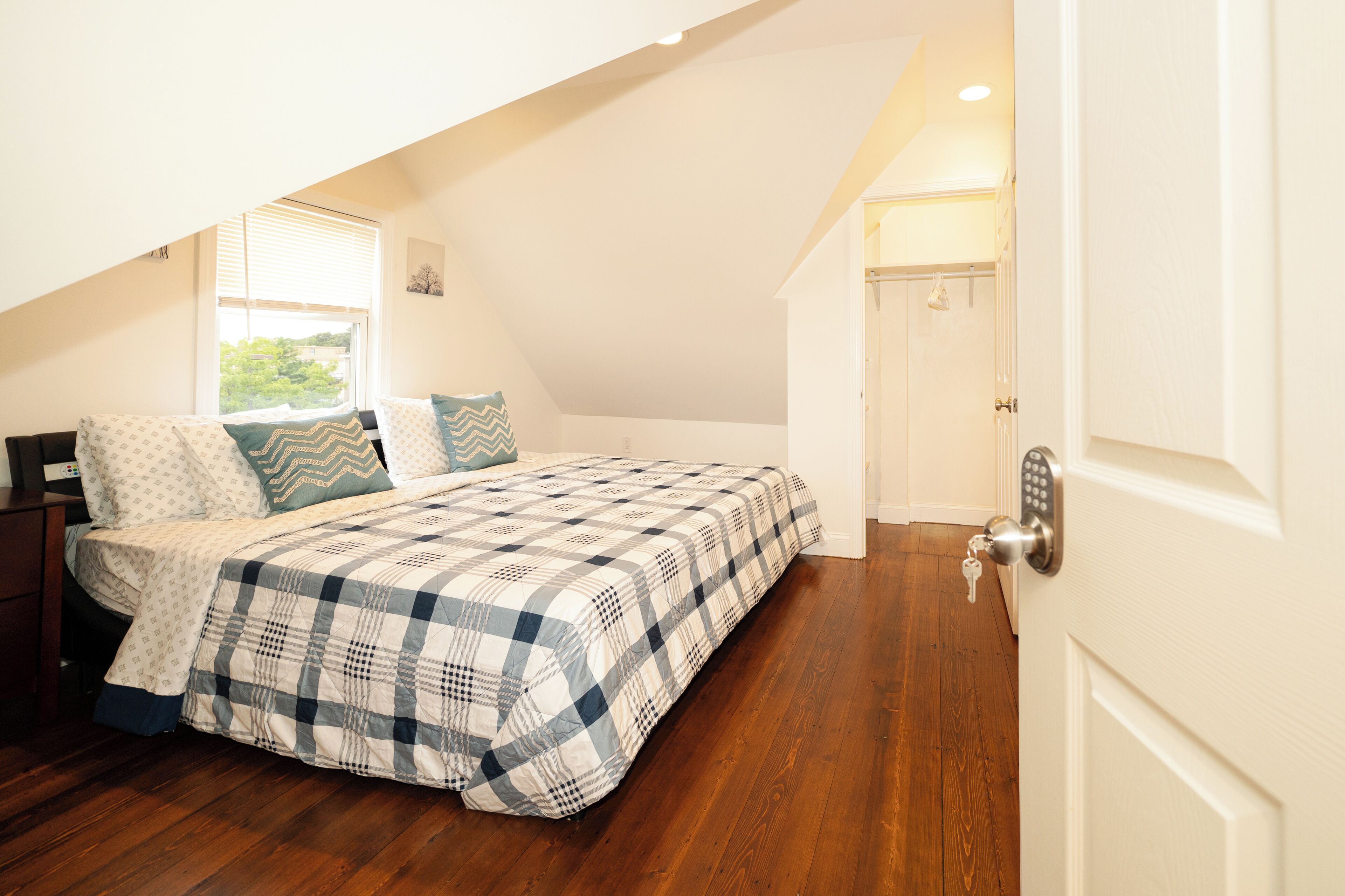 Une chambre avec une couvre-lit à carreaux, un plancher en bois, une table de chevet et une porte dotée d’un système de verrouillage à clavier.