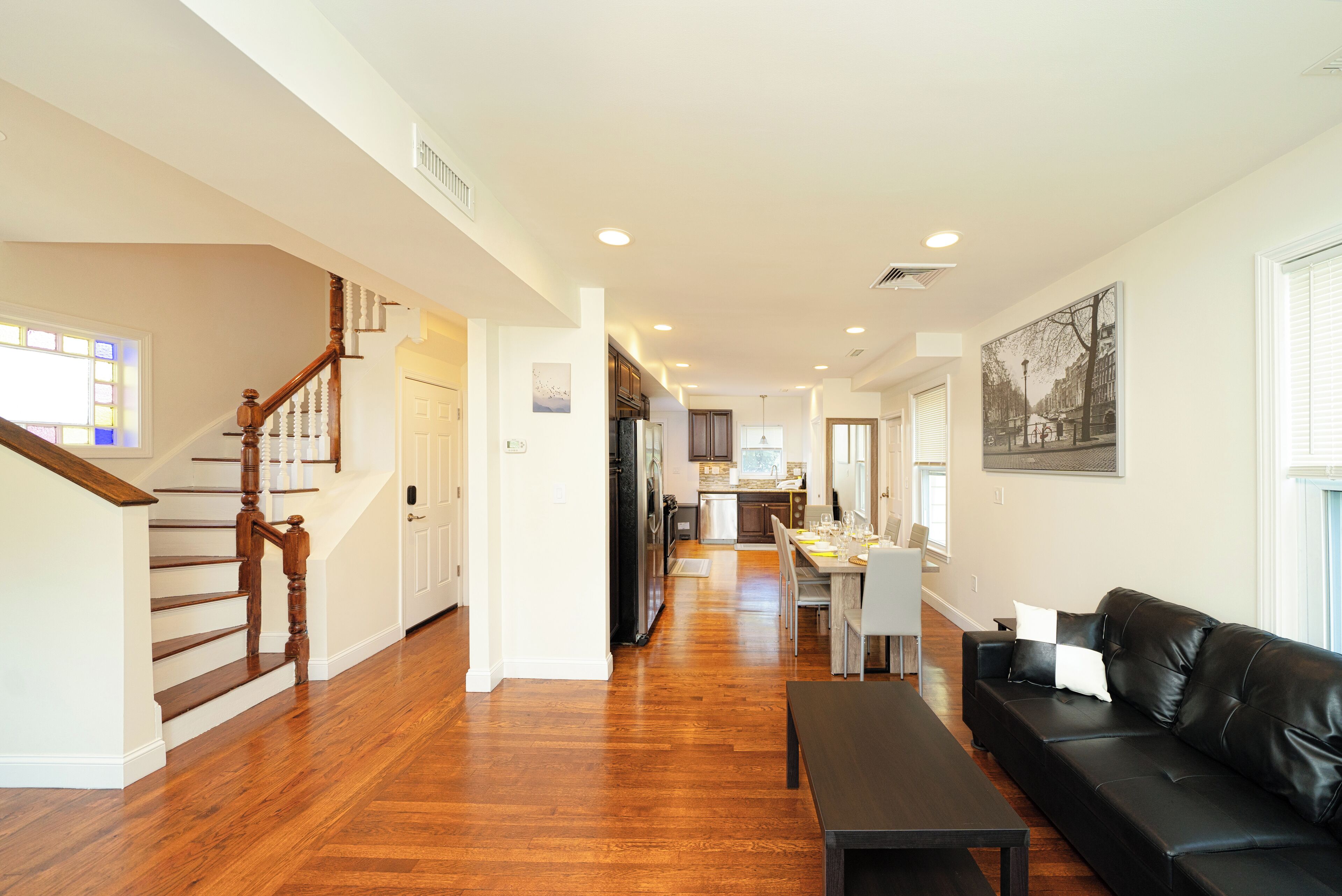 A spacious living area with hardwood flooring, a black leather sofa, a dining table, and a kitchen in the background.