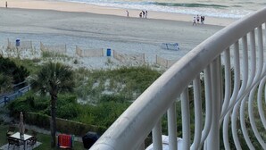 Vista de la playa desde un balcón con barandilla, olas del océano y gente caminando sobre la arena.