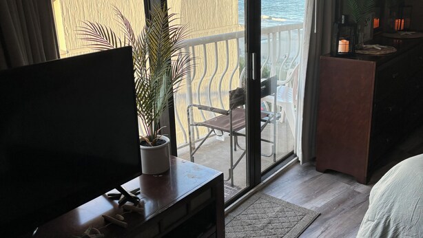Habitación de hotel con un amplio ventanal corredero que da a un balcón con vistas al mar. Hay un televisor sobre un mueble de madera, una pequeña planta en maceta y una mesita de noche con una lámpara.