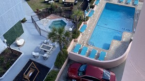 Zona de piscina con sillones de descanso y una bañera de hidromasaje, un coche rojo aparcado cerca y vistas a la playa con gente y sombrillas.