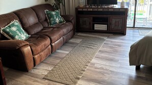 Una sala de estar con un sofá de cuero marrón, un televisor sobre un mueble de madera, un ventanal con cortinas y una planta.