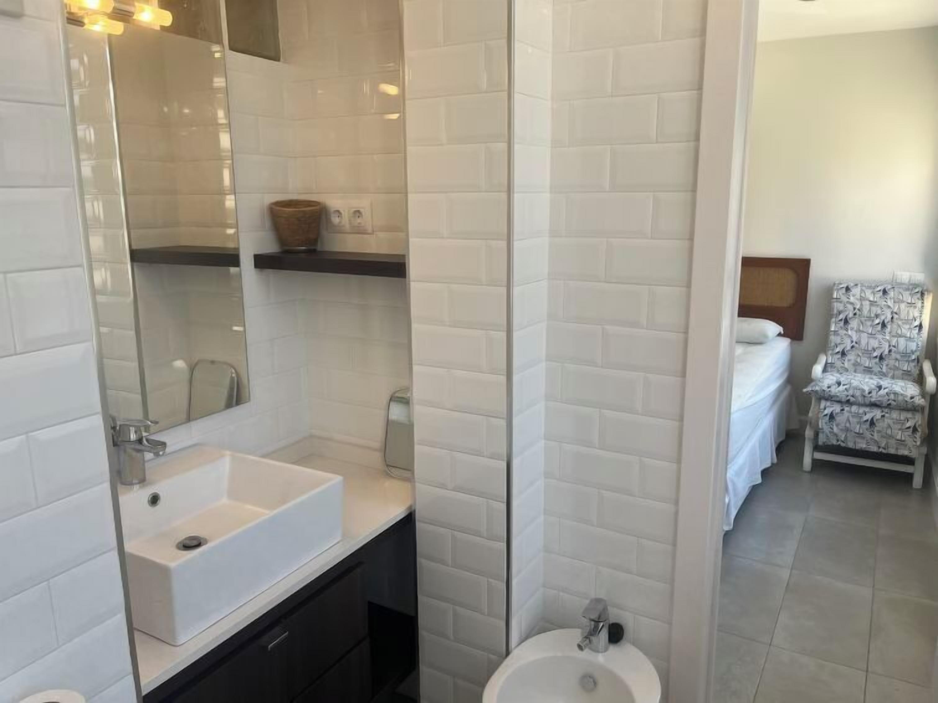 Une salle de bain avec un lavabo blanc, un miroir et des toilettes. Dans la pièce adjacente, on trouve un lit et une chaise  à motifs.