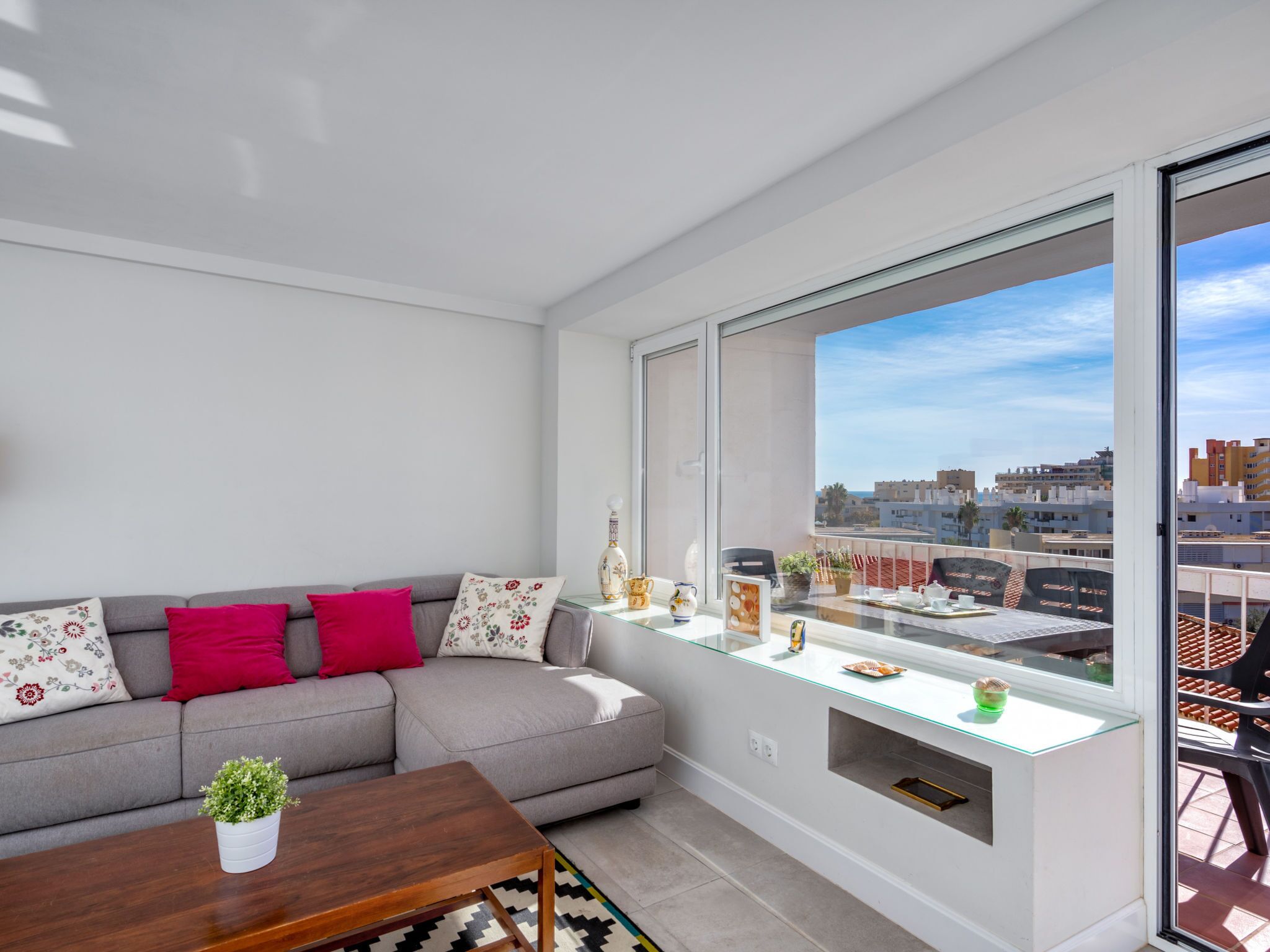 A modern apartment with a grey sofa, red pillows, a wooden coffee table, and a balcony with a city view.