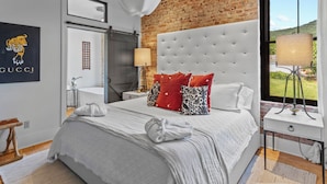 A bedroom with a large bed, a tufted headboard, a brick wall, and a view of the outdoors. - Top of the Town | Luxury 2BR Adults-Only · Downtown Burnsville · Walk Everywhere (Burnsville)