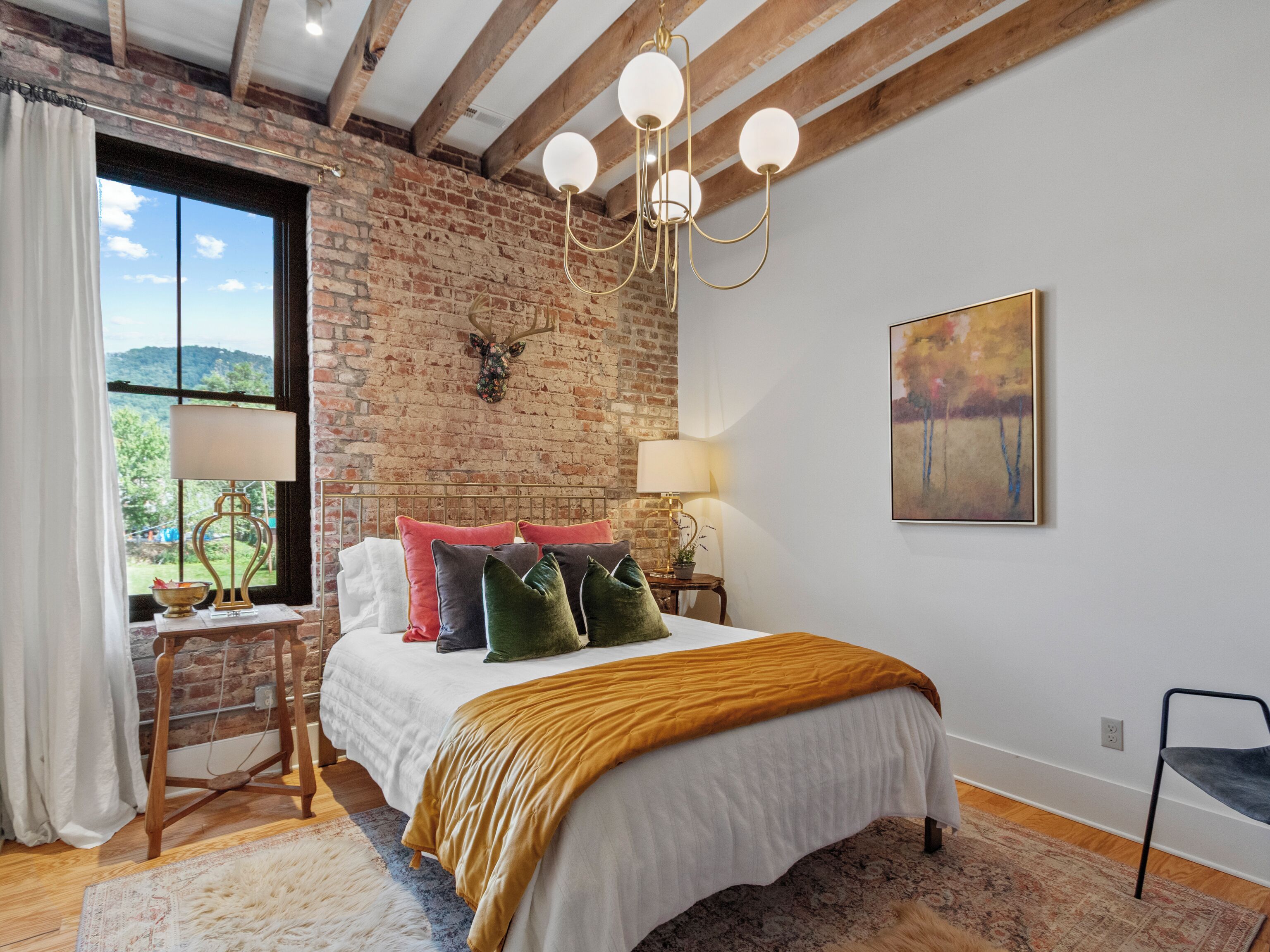 Ein Schlafzimmer mit Backsteinwand, einem Bett mit vielen Kissen, einer Chandelieber, einem Gemälde und Blick durch ein Fenster.