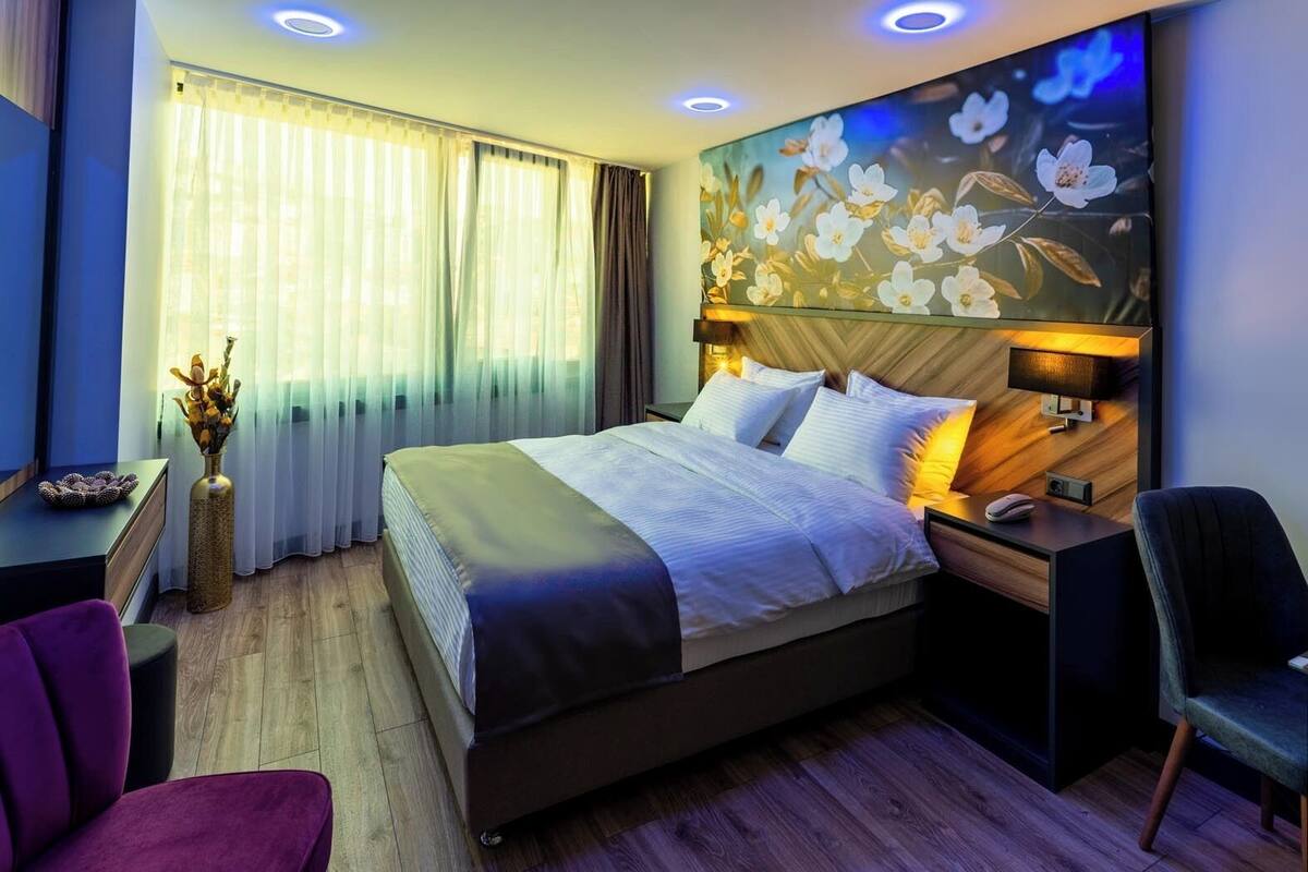 Ett hotellrum med en stor säng, ett skrivbord, en stol, en vas med blommor och en vägg prydd med en blomstermålning.