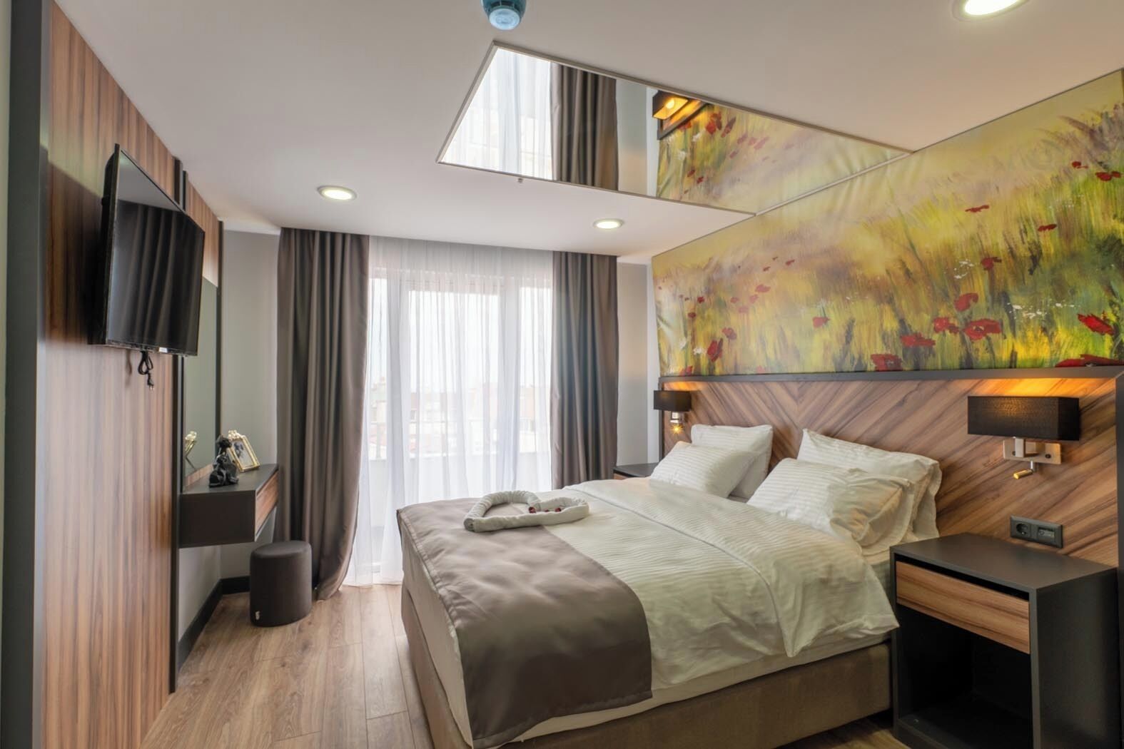 A modern hotel room with a large bed, a flat-screen TV mounted on the wall, a large abstract painting, and a window with sheer curtains.