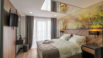 A modern hotel room with a large bed, a flat-screen TV mounted on the wall, a large abstract painting, and a window with sheer curtains.