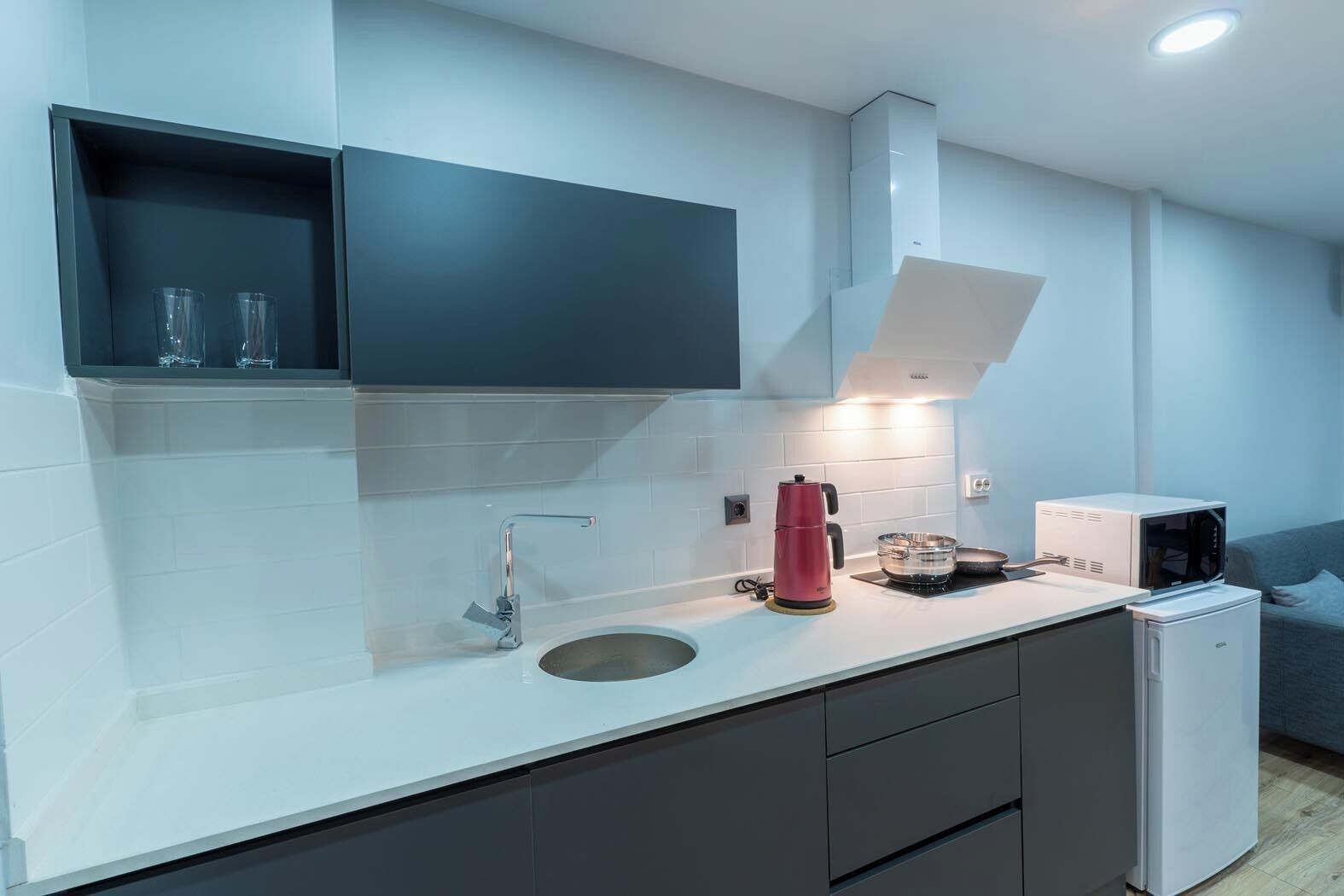 A modern kitchen with a white countertop, dark cabinets, a stainless steel sink, a red kettle, a microwave, and a small refrigerator.