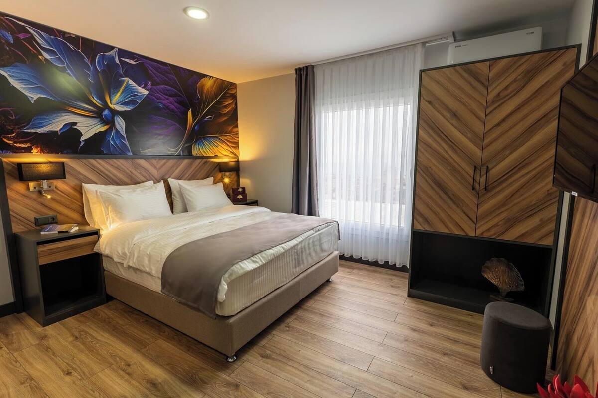 Ett modernt hotellrum med en stor säng, ett träfärgat sänggavel, en väggmonterad TV och ett stort fönster med tunna, genomskinliga gardiner.
