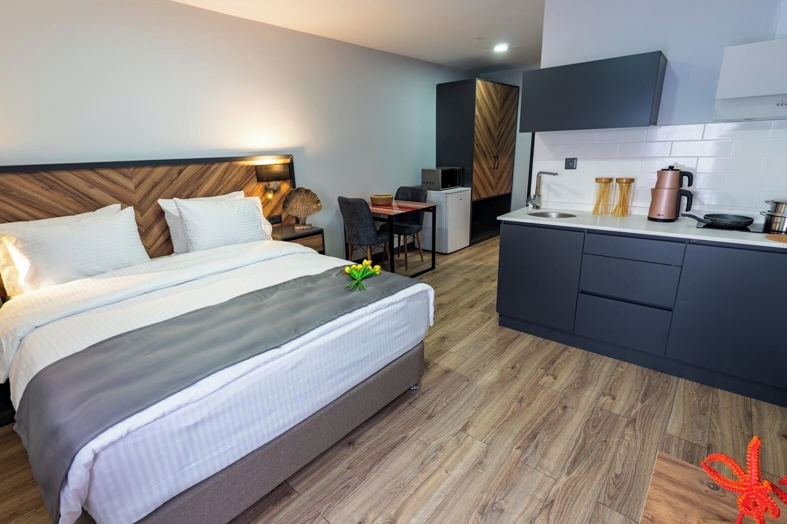 A hotel room with a bed, bedside table, desk, chair, kitchenette, and a small dining area.