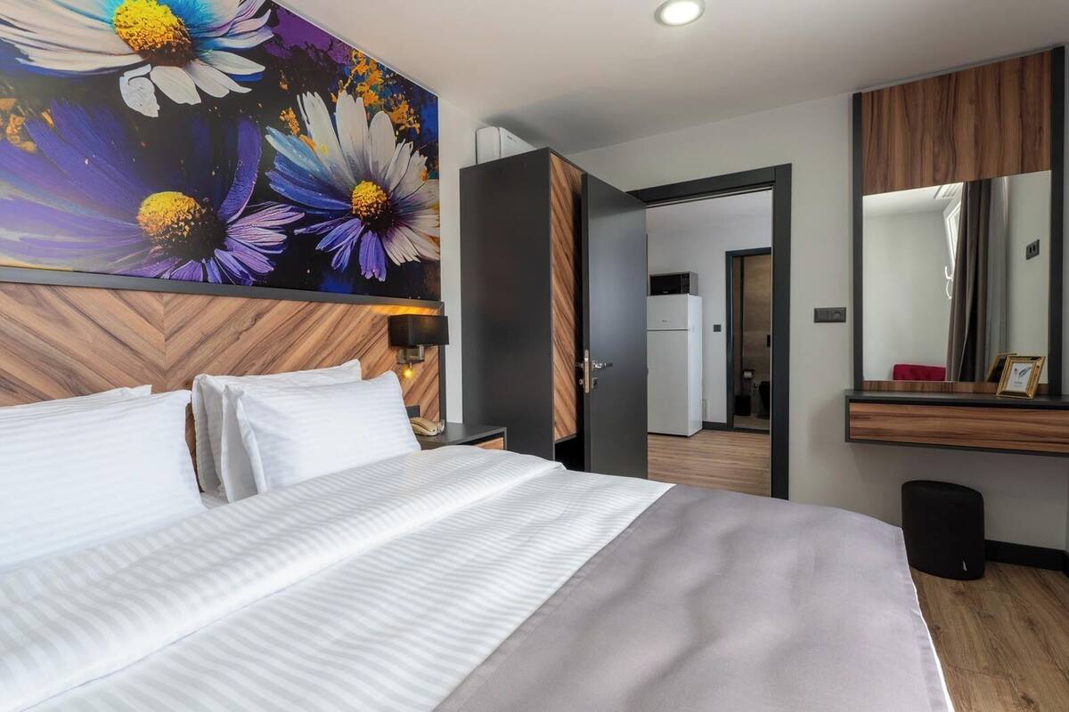 Ett modernt hotellrum med en stor säng, en träpanel som huvudgavel, en tavla med blommor, en spegel och utsikt in i badrummet.