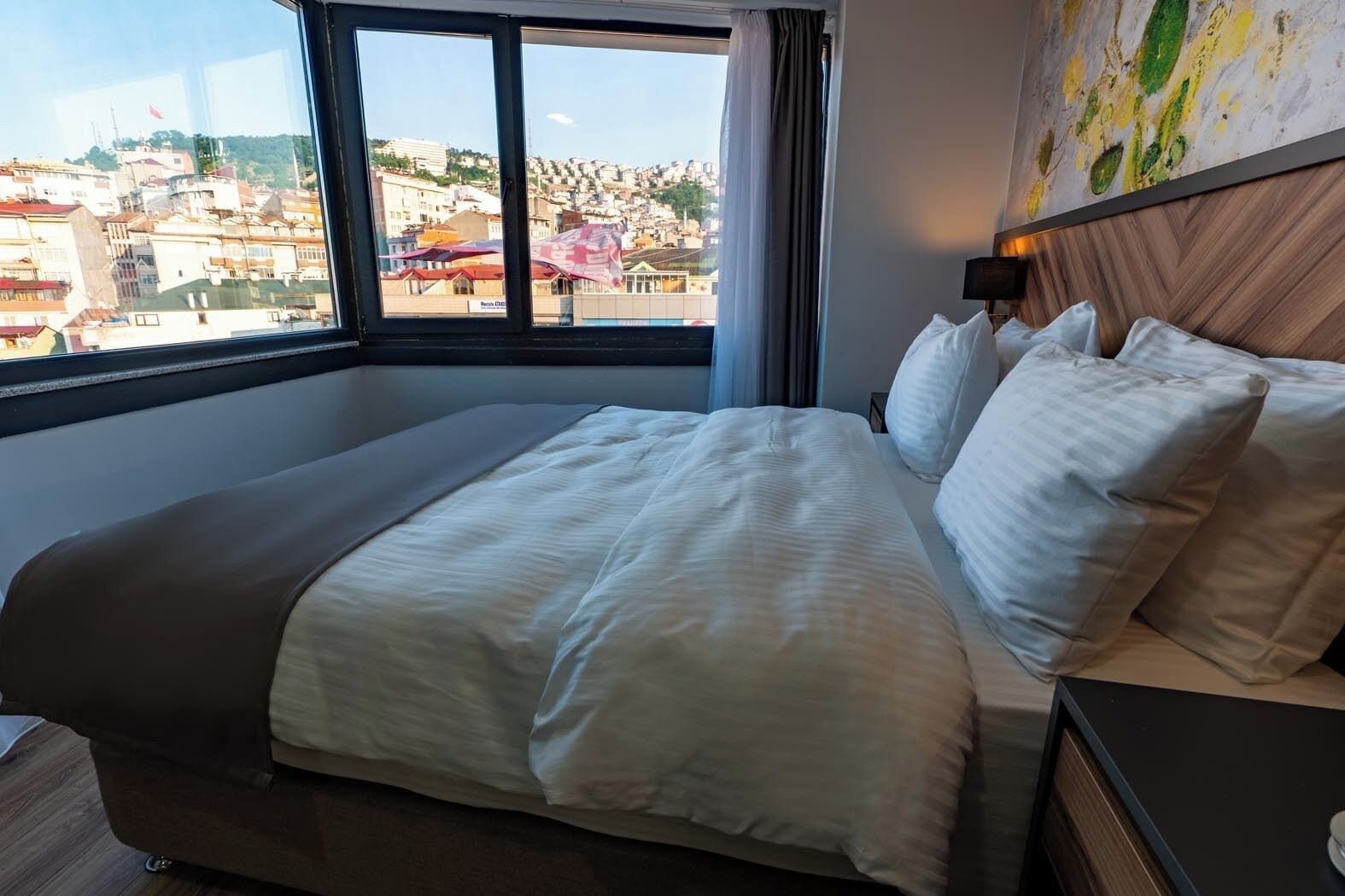 A hotel room with a large bed, a nightstand, a lamp, and a view of a cityscape through a window.