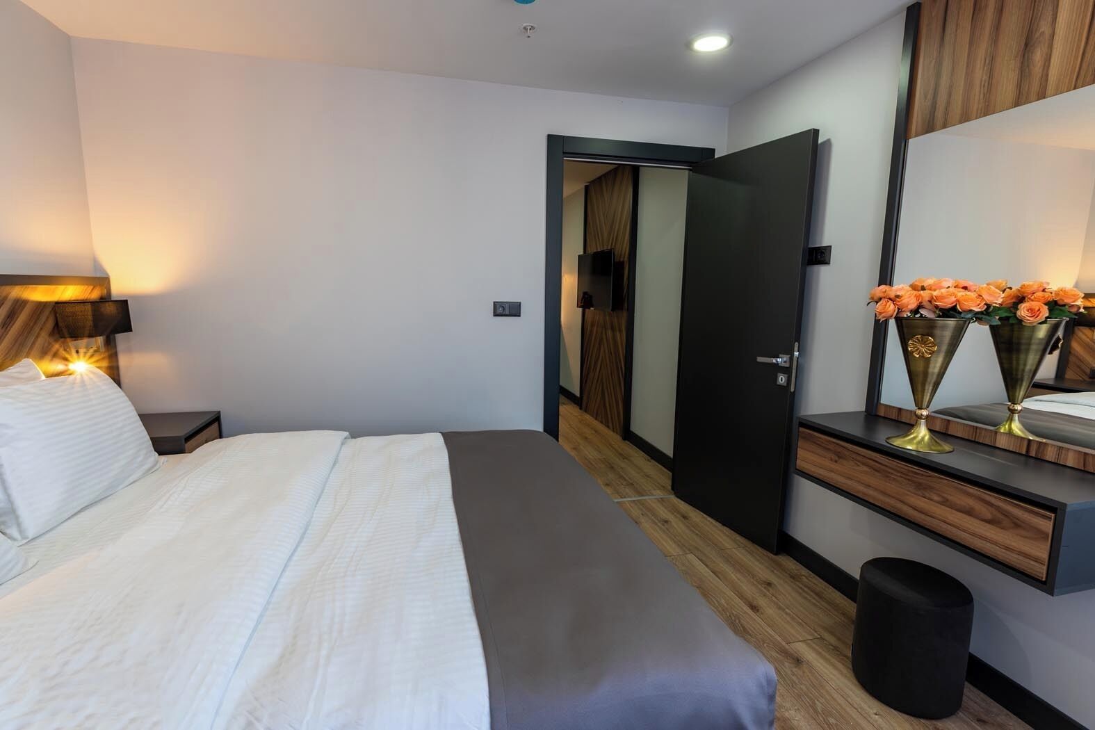 A modern hotel room with a bed, bedside table, lamp, mirror, and a small trash bin.