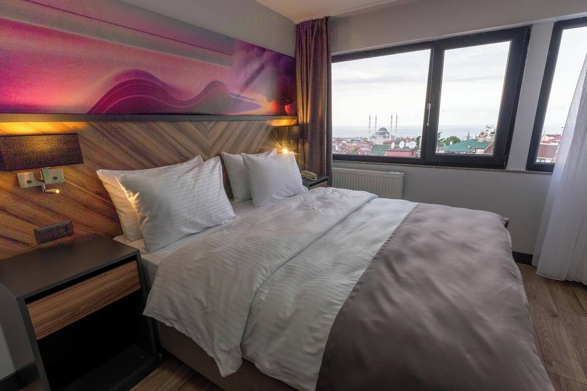 Ett hotellrum med en säng, sängbord, en vägglampa och ett fönster med utsikt över stadsbilden.