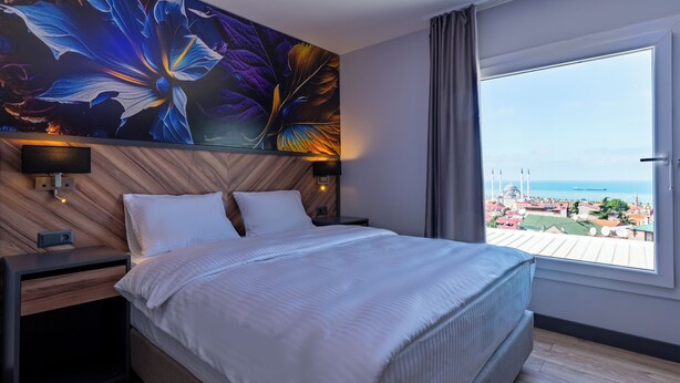 Una habitación de hotel moderna con una cama grande, dos mesitas de noche, un televisor montado en la pared y un ventanal con vistas a la ciudad.