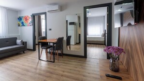 Una habitación de hotel moderna con suelo de madera, un sofá gris, una mesa de comedor con sillas, un espejo, una cama con edredón blanco y un jarrón con flores.