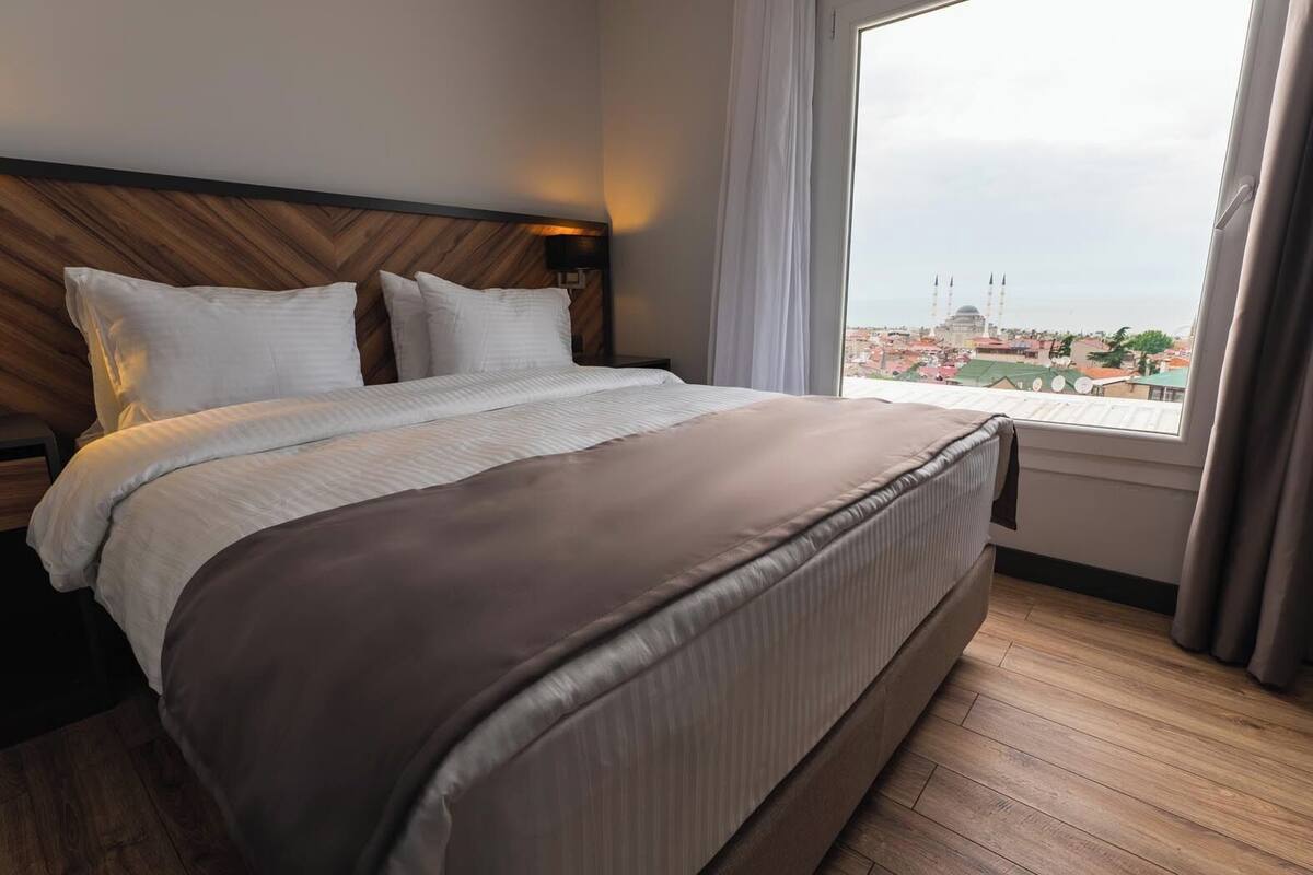 Ett hotellrum med en stor säng, en sänggavel i trä och utsikt över en stadssiluett genom ett fönster.