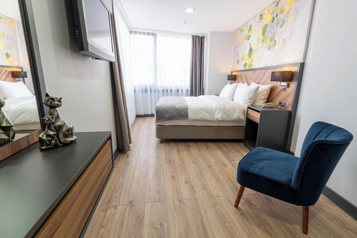 Ett modernt hotellrum med en säng, ett skrivbord, en stol, en TV och ett stort fönster med gardiner.
