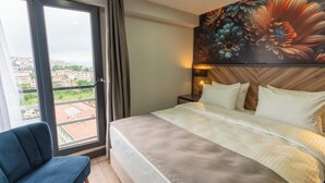 Habitación de hotel con cama, una silla, un ventanal con vistas a edificios y un mural floral en la pared.