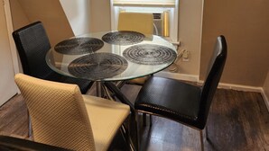 A dining area with a glass table and black chairs, a window with blinds, and a beige wall.