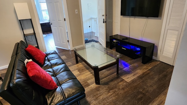 A living room with a black leather sofa, a glass coffee table, a flat-screen TV mounted on the wall, and a bench with blue LED lighting.