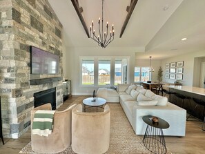 A spacious living room with a stone fireplace, a large sofa, a coffee table, and a dining area with a bar. - Modern Farmhouse - 4 Bd 3 Bth (Lincoln)