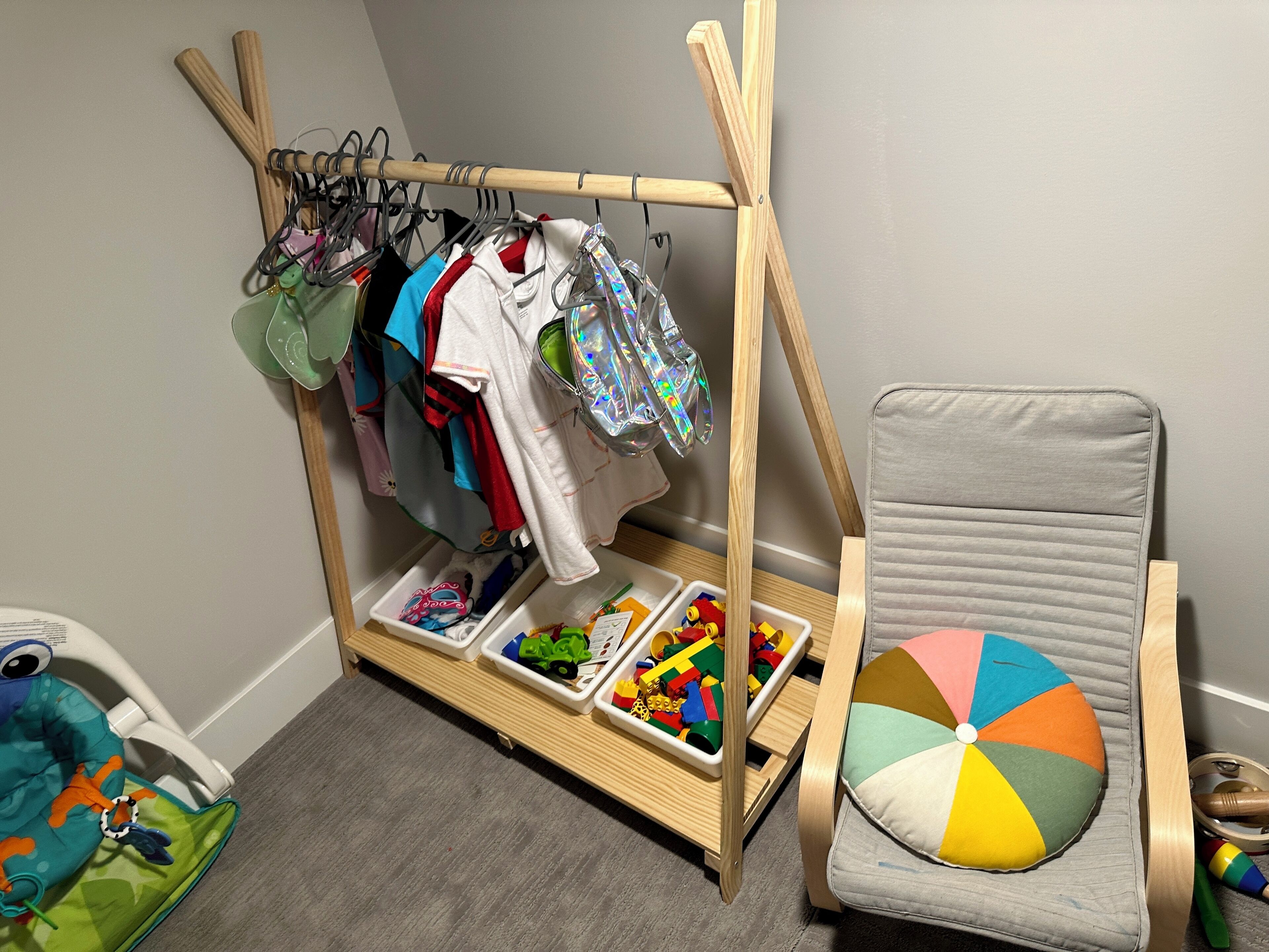 A child's play area with a wooden rack holding clothes, a chair with a colorful cushion, and storage bins with toys.