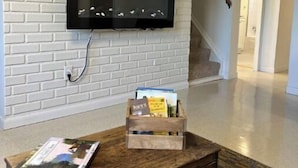 A living room with a white brick wall, a mounted flat-screen TV, a wooden coffee table with magazines, and a staircase in the background. - The Red House -  modern beautiful Scandinavian style cozy cottage! (Chassell)