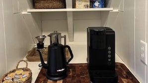 A kitchen area with white shelves holding white mugs, a coffee maker, a kettle, and a basket of coffee pods. - The Red House -  modern beautiful Scandinavian style cozy cottage! (Chassell)