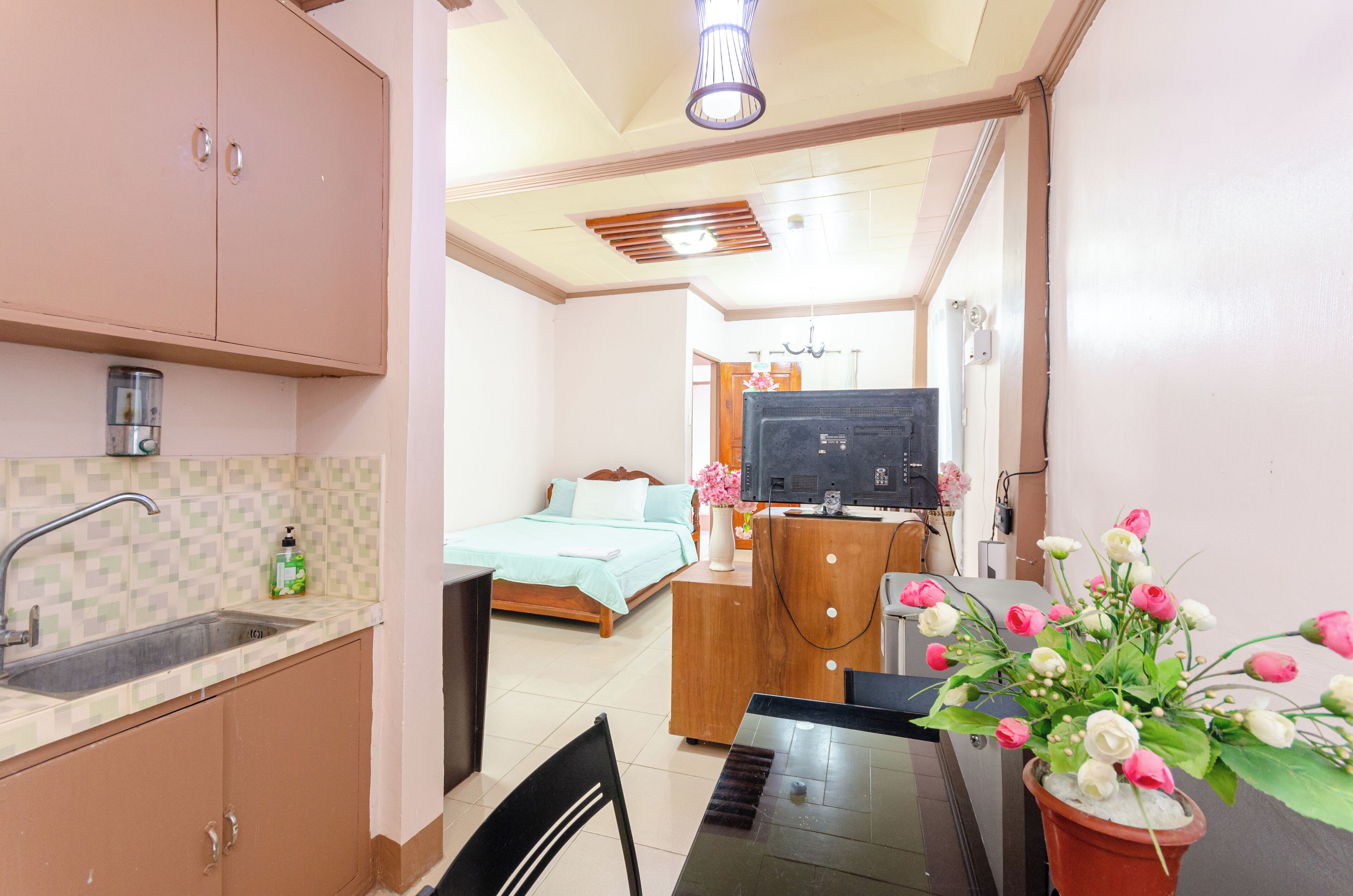 A compact hotel room with a kitchenette, a bed with a blue bedspread, a wooden dresser, a TV, and a potted plant with flowers.