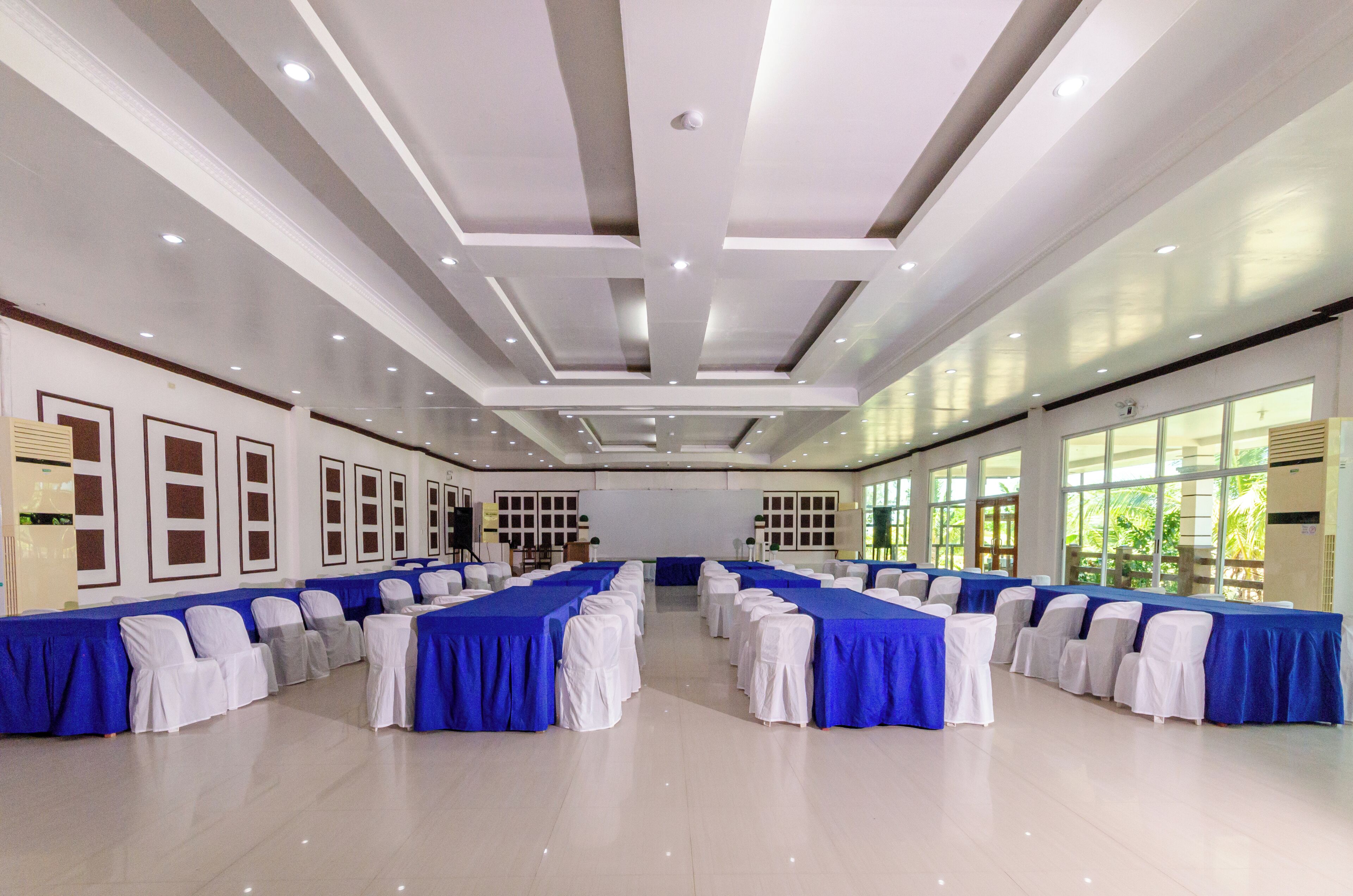 A large, well-lit conference room with rows of tables and chairs, large windows, and a modern ceiling design.
