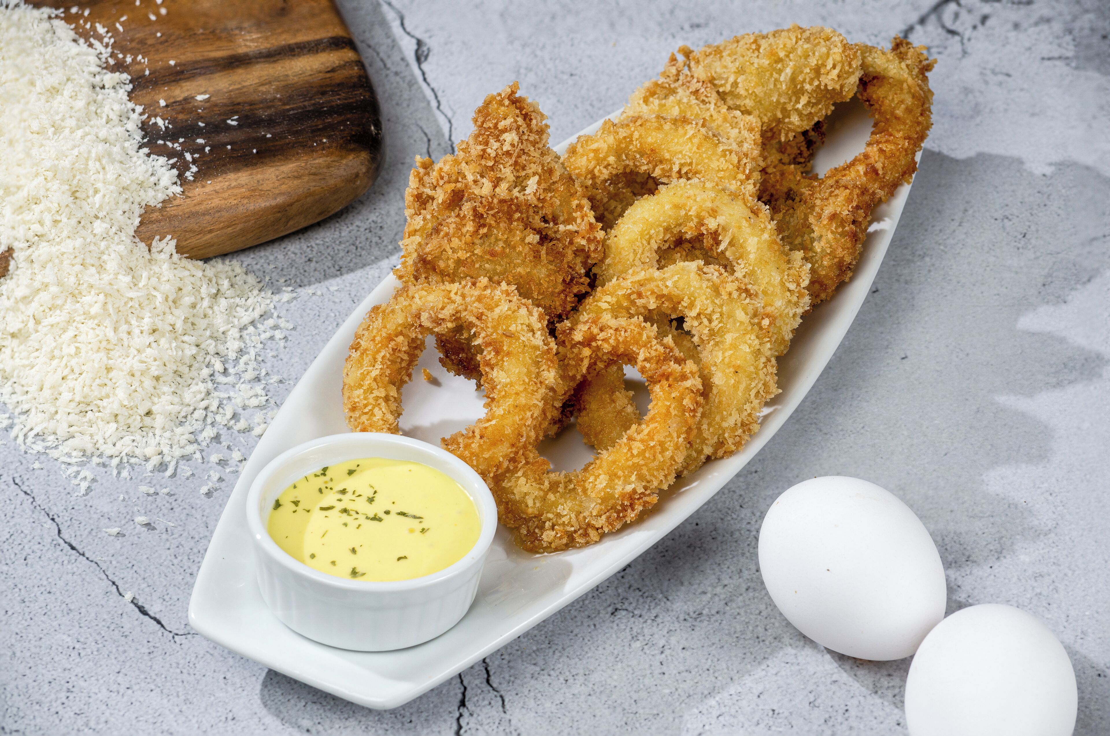 Un plato de cebollas a la romana crujientes con salsa para mojar y dos huevos sobre una superficie rugosa.