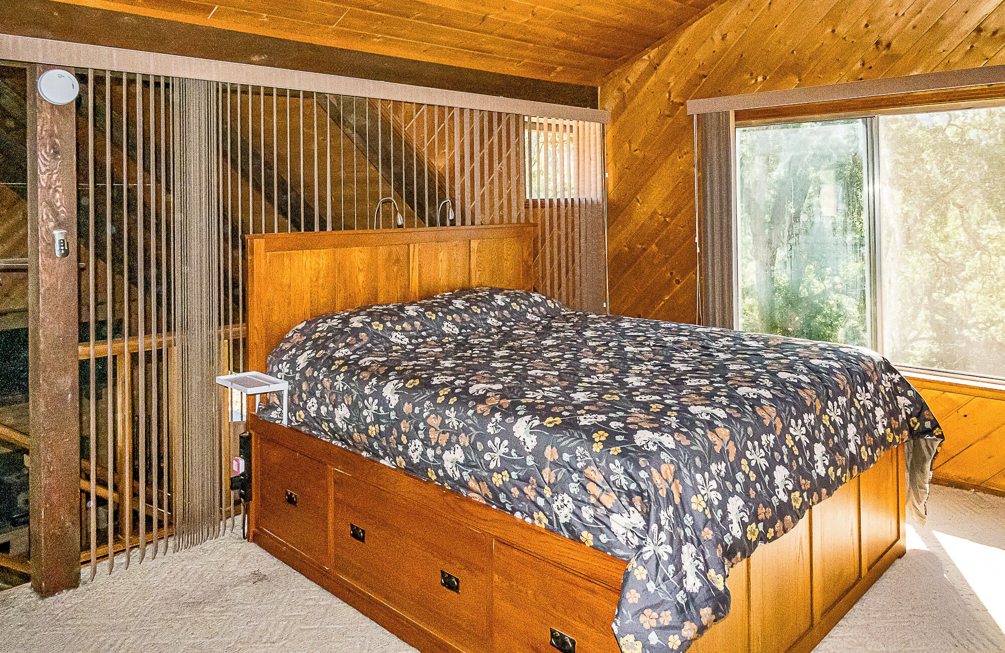 A wooden-framed bed with a floral bedspread in a room with a large window and a view of trees.