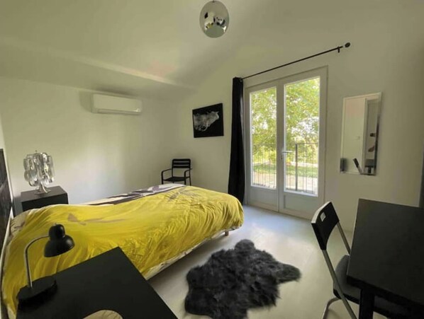 Une chambre avec un lit, un bureau, une chaise, un tapis, un tableau au mur et un balcon donnant sur un paysage verdoyant.