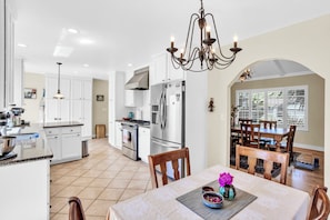 A modern kitchen with white cabinets, stainless steel appliances, and a dining area with wooden chairs and a chandelier.