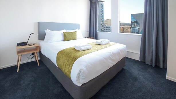 A hotel room with a large bed, a bedside table with a lamp, and a view of a cityscape through the window. - Liberty Apartment Hotel (Wellington)