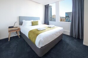 A hotel room with a large bed, a bedside table with a lamp, and a view of a cityscape through the window. - Liberty Apartment Hotel (Wellington)