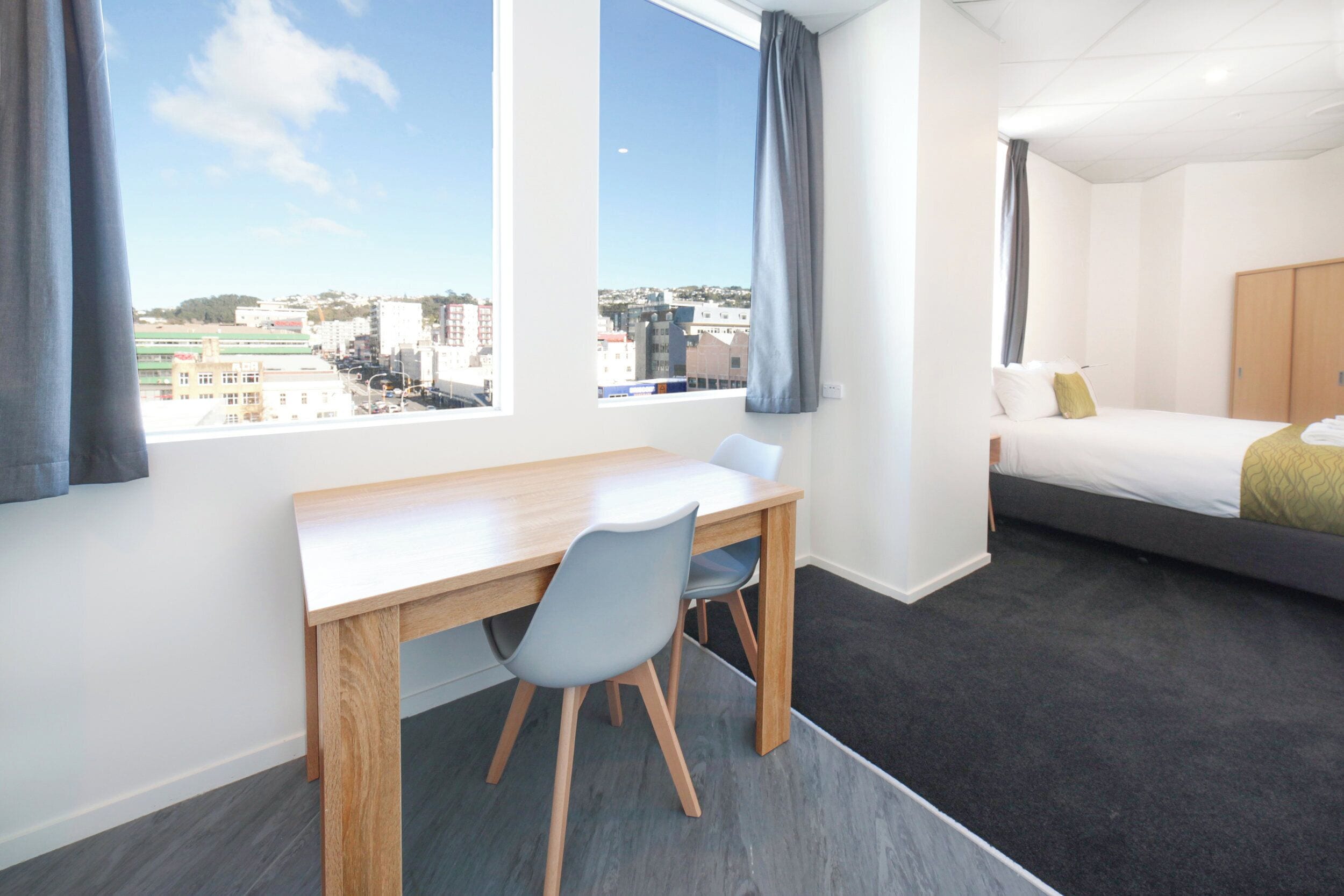 A modern hotel room with a wooden desk, two chairs, a bed with a grey mattress and a yellow throw, and a large window offering a city view.