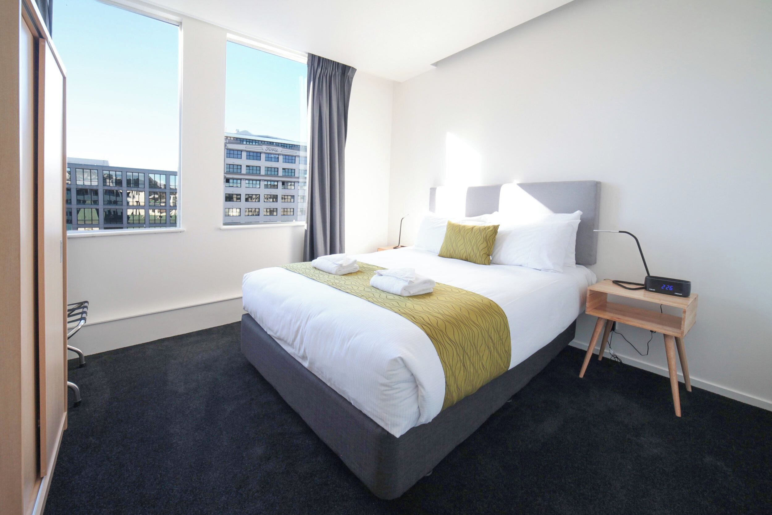 Una habitación de hotel con una cama grande, una mesita de noche con una radio y vistas a edificios a través de la ventana.