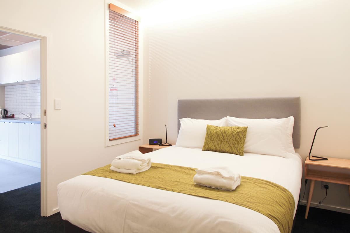 Une chambre d’hôtel comprenant un lit, une table de chevet, une commode, une fenêtre avec des stores et une vue sur la cuisine.