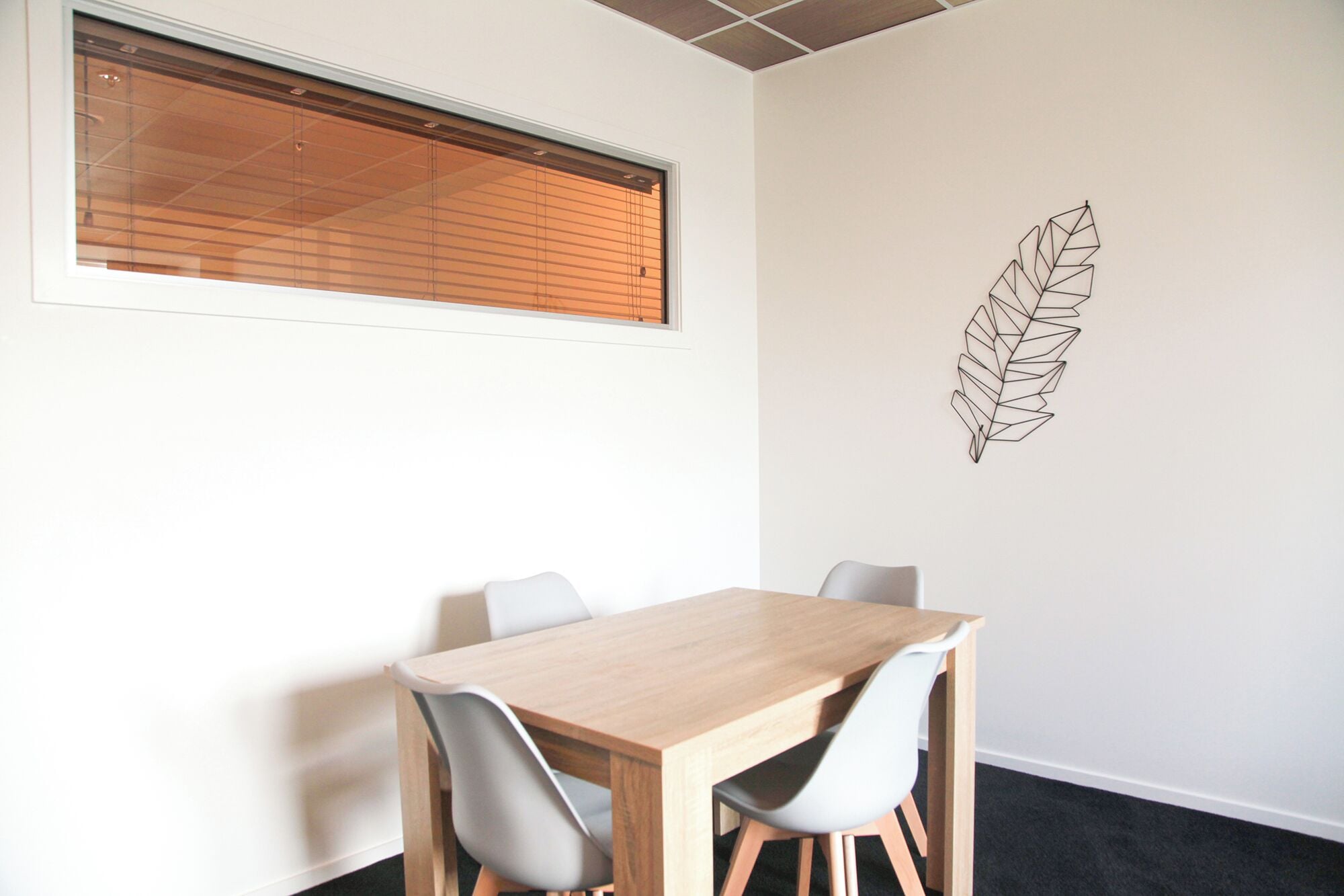 Une salle de conférence avec une table en bois et quatre chaises, une fenêtre avec des stores et une décoration murale représentant des feuilles.