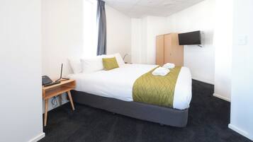 A hotel room with a large bed, a desk with a computer, a TV mounted on the wall, and a wardrobe.
