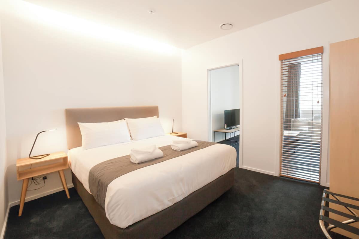 Une chambre d’hôtel moderne avec un grand lit, des tables de chevet, une télévision et une fenêtre équipée de stores.
