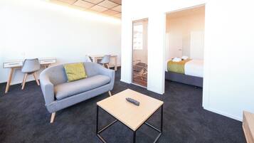 Quarto de hotel moderno com um sofá cinza, uma pequena mesa de madeira com um controle remoto, uma escrivaninha com uma cadeira e uma cama com um cobertor verde.