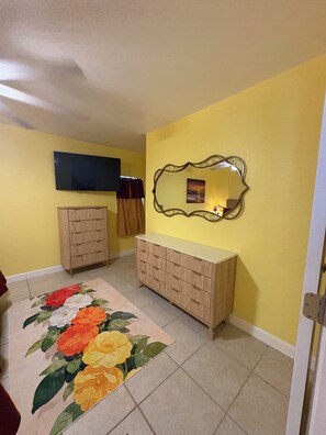 A room with a yellow wall, a wooden cabinet, a flat-screen TV, a decorative mirror, and a floral rug.