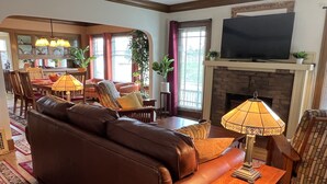 Ein Wohnzimmer mit braunem Ledersofa, einem Holztisch, einem Fernseher über einem Kamin und einem Essbereich mit Tisch und Stühlen.