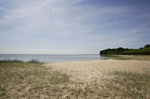 Une plage de sable avec quelques touffes d’herbe et une mer calme sous un ciel dégagé.
