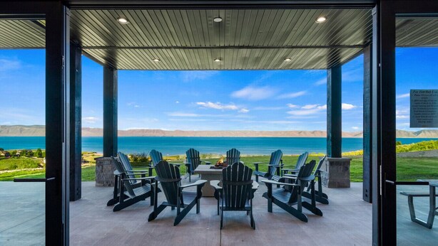 A covered outdoor seating area with Adirondack chairs and a table, overlooking a body of water and distant hills.